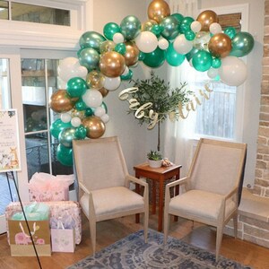 Op de afbeelding: Een groene, gouden en witte ballonboog met de woorden "baby claire" in gouden glitterletters. Twee beige stoelen staan voor de boog, en een kleine tafel met een plant in een pot staat ertussen.