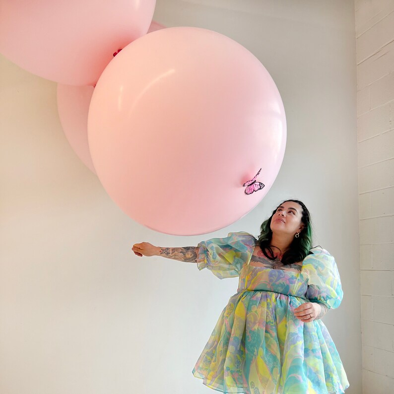 Puede incluir: Una mujer con un vestido floral con un gran globo rosa sobre su cabeza. El globo est&aacute; decorado con una mariposa rosa y la mujer lo est&aacute; mirando.