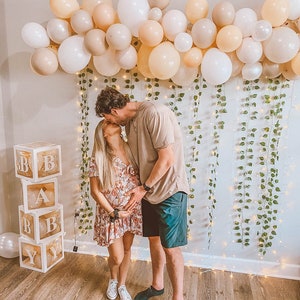 Może przedstawiać: Para całuje się przed ścianą ozdobioną zielenią i światełkami. Para stoi przed stosem przezroczystych pudełek z literami "BABY" napisanymi na nich.