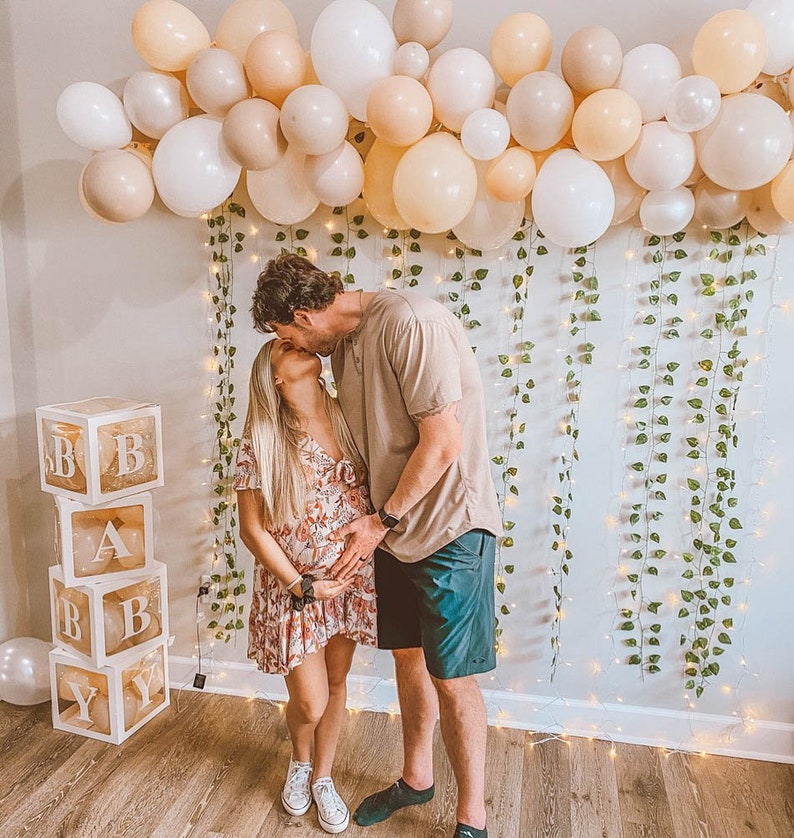 Może przedstawiać: Para całuje się przed ścianą ozdobioną zielenią i światełkami. Para stoi przed stosem przezroczystych pudełek z literami "BABY" napisanymi na nich.