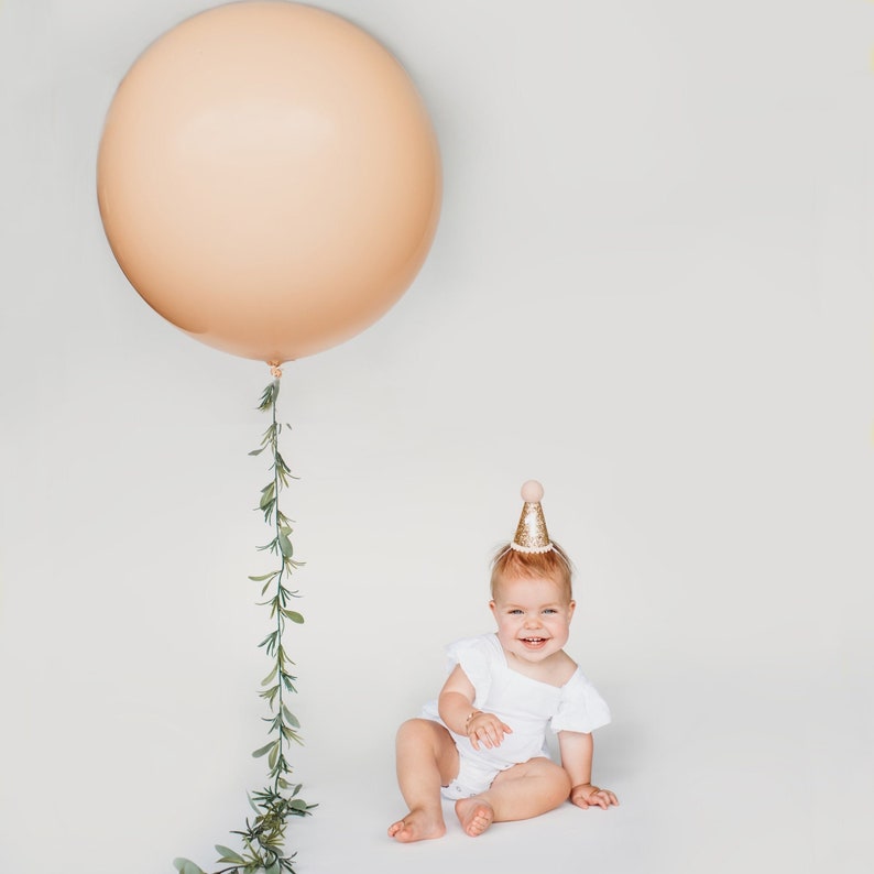 May include: A baby wearing a white dress and a gold glitter party hat sits on a white background. A large peach balloon with a green garland hangs above the baby.