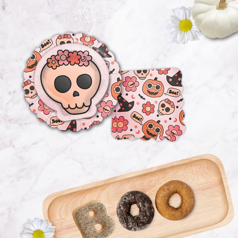 May include: Pink and white paper plates with a skull and crossbones design, a pink and white paper napkin with a skull and crossbones design, and a wooden tray with three donuts. The donuts are a chocolate donut, a glazed donut, and a donut with sprinkles.