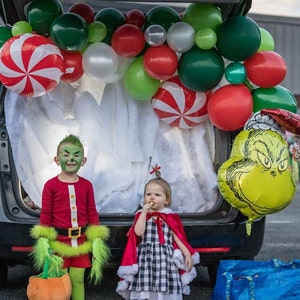 Pu&ograve; includere: Due bambini vestiti in costume per Halloween. La bambina &egrave; vestita come Cindy Lou Who dal film Il Grinch. Indossa un vestito a scacchi rosso e bianco con un mantello rosso. Il bambino &egrave; vestito come il Grinch. Indossa un costume verde e rosso con un cappello verde. Sono in piedi davanti a un'auto decorata con palloncini di Natale.