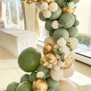 Puede incluir: Un arco de globos verde y crema con flores blancas y vegetaci&oacute;n. Los globos est&aacute;n dispuestos en cascada, creando un tel&oacute;n de fondo festivo y elegante.