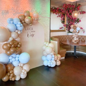 Op de afbeelding: Een beige achtergrond met de tekst "Luca turns One" in bruin schrift. De achtergrond is versierd met een ballonboog gemaakt van bruine, beige en lichtblauwe ballonnen.