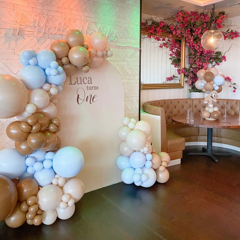 Op de afbeelding: Een beige achtergrond met de tekst "Luca turns One" in bruin schrift. De achtergrond is versierd met een ballonboog gemaakt van bruine, beige en lichtblauwe ballonnen.