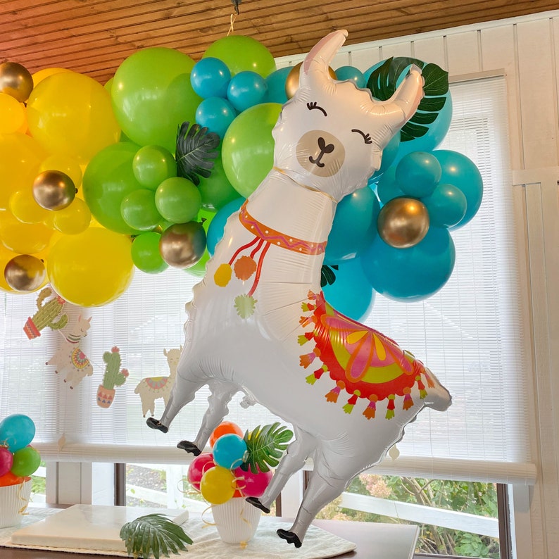 May include: A white llama balloon with a colorful patterned saddle, surrounded by yellow, green, and blue balloons. The llama balloon has a friendly face with closed eyes and a small nose.