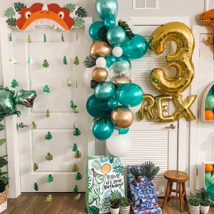 Op de afbeelding: Een groene en gouden dinosaurus-thema verjaardagsdecoratie met een ballonboog, een grote gouden ballon met het nummer 3 en het woord "REX" in gouden letters. De ballonboog is gemaakt van groene, gouden en witte ballonnen. Er is een groene dinosaurus-uitsparing op de muur en een groene dinosaurus-uitsparing op een cadeauzak.
