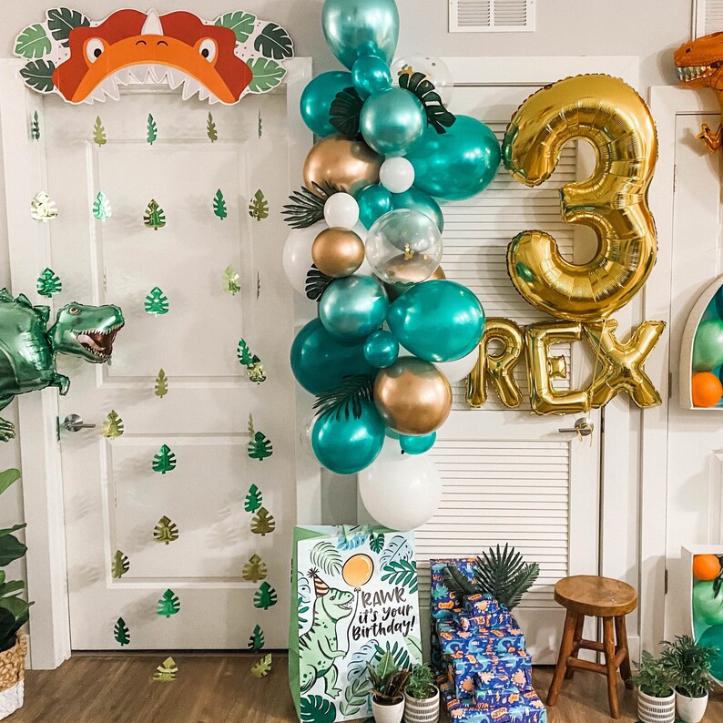 Op de afbeelding: Een groene en gouden dinosaurus-thema verjaardagsdecoratie met een ballonboog, een grote gouden ballon met het nummer 3 en het woord "REX" in gouden letters. De ballonboog is gemaakt van groene, gouden en witte ballonnen. Er is een groene dinosaurus-uitsparing op de muur en een groene dinosaurus-uitsparing op een cadeauzak.