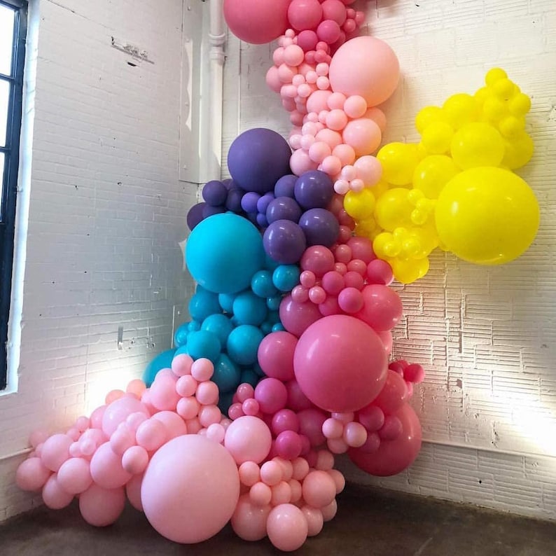 Op de afbeelding: Een muur versierd met een kleurrijke ballonversiering. De versiering bestaat uit roze, paarse, blauwe en gele ballons in verschillende maten. De ballonnen zijn in een watervalpatroon gerangschikt, wat een feestelijke en speelse uitstraling cre&euml;ert.