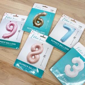 May include: A set of 32-inch number balloons in pink, gold, blue, and white. The balloons are packaged in individual clear plastic bags with a blue and white label that says "Number Balloon" and "Ellie's Brand".