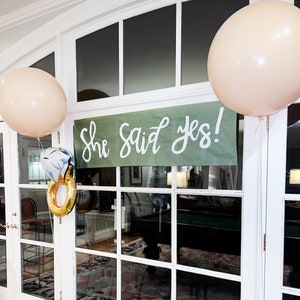 K&ouml;nnte beinhalten: Ein gr&uuml;nes Banner mit dem Text "She said yes!" h&auml;ngt in einem Fenster. Im Hintergrund sind zwei beigefarbene Ballons zu sehen.