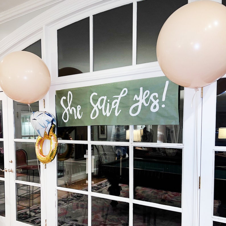 K&ouml;nnte beinhalten: Ein gr&uuml;nes Banner mit dem Text "She said yes!" h&auml;ngt in einem Fenster. Im Hintergrund sind zwei beigefarbene Ballons zu sehen.