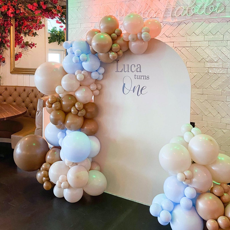 Op de afbeelding: Een witte boog achtergrond versierd met een ballon slinger in tinten bruin, beige en blauw. De slinger is gemaakt van verschillende maten ballonnen. De achtergrond heeft de tekst "Luca turns One" erop.