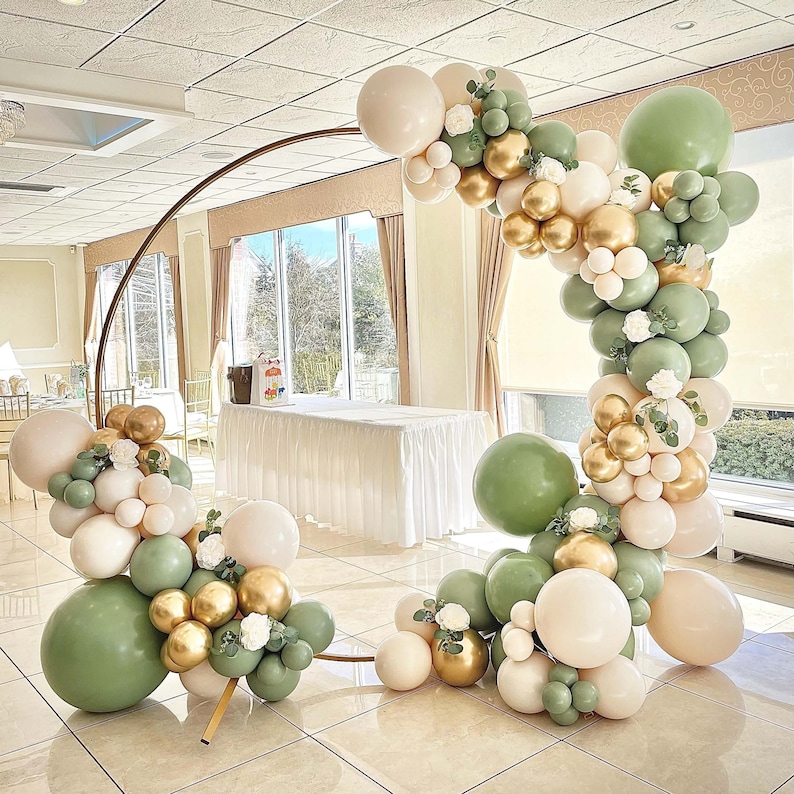 Puede incluir: Un arco de metal dorado decorado con una guirnalda de globos verdes, blancos y dorados. Los globos est&aacute;n dispuestos en cascada, con los globos verdes en la parte superior y los globos blancos en la parte inferior. El arco est&aacute; instalado en una habitaci&oacute;n con paredes blancas y un mantel blanco sobre una mesa.