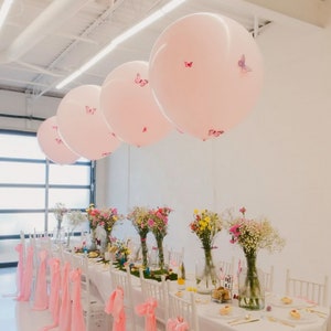 Puede incluir: Una escena de fiesta con grandes globos rosas adornados con calcomanías de mariposas suspendidos sobre una larga mesa. La mesa está puesta con manteles blancos, centros de mesa florales y cintas rosas.