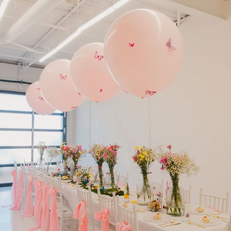 Puede incluir: Una mesa puesta con sillas blancas y un mantel blanco. Hay globos rosas con decoraciones de mariposas colgando del techo. La mesa est&aacute; decorada con centros de mesa florales en jarrones de vidrio.