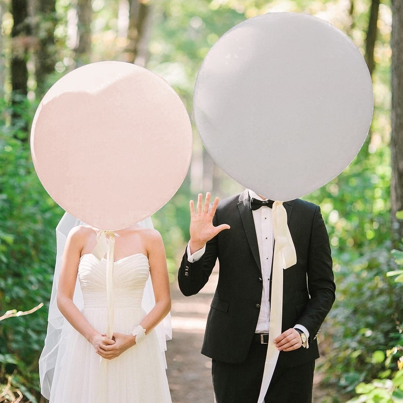 May include: A bride and groom holding large pink and gray balloons in front of their faces. The bride is wearing a white wedding dress and the groom is wearing a black suit and a white bow tie.