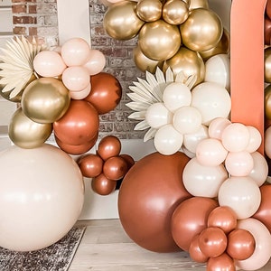 K&ouml;nnte beinhalten: Eine Sammlung von Ballons in verschiedenen Braunt&ouml;nen, Beige und Gold. Die Ballons sind in Clustern angeordnet und mit einer Schnur zusammengebunden. Die Ballons befinden sich vor einem Hintergrund aus Backsteinmauer.