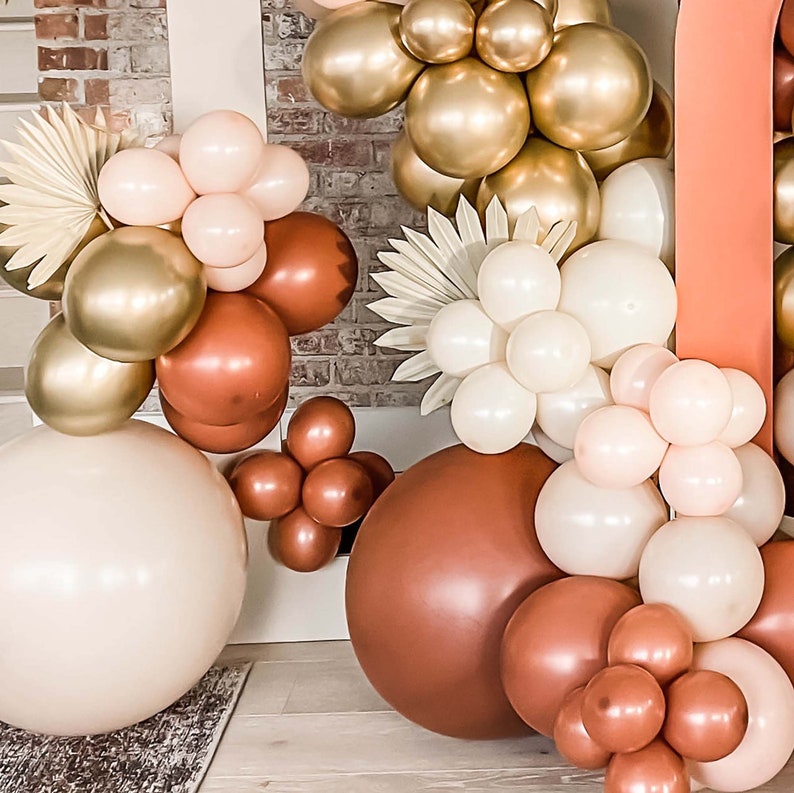 K&ouml;nnte beinhalten: Eine Sammlung von Ballons in verschiedenen Braunt&ouml;nen, Beige und Gold. Die Ballons sind in Clustern angeordnet und mit einer Schnur zusammengebunden. Die Ballons befinden sich vor einem Hintergrund aus Backsteinmauer.