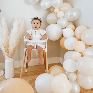 May include: A baby girl wearing a white dress and a floral headband sits in a high chair. The background is decorated with a garland of white and beige balloons.