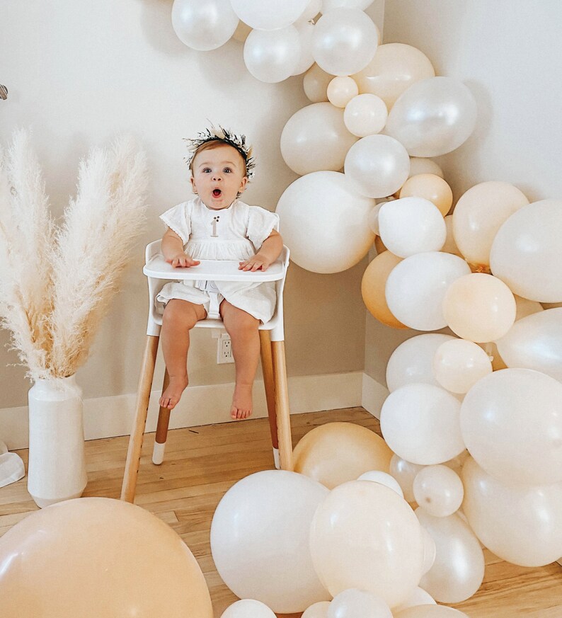 May include: A baby girl wearing a white dress and a floral headband sits in a high chair. The background is decorated with a garland of white and beige balloons.