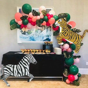 May include: A black and white zebra balloon and a gold tiger balloon with green and pink balloons in the background. The balloons are arranged in an arch shape and are decorated with green leaves. There is a black table with a white tablecloth and a variety of snacks on it.