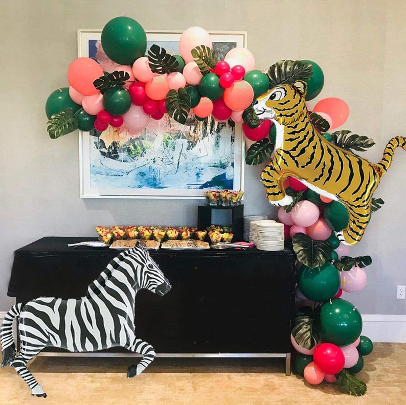 May include: A black and white zebra balloon and a gold tiger balloon with green and pink balloons in the background. The balloons are arranged in an arch shape and are decorated with green leaves. There is a black table with a white tablecloth and a variety of snacks on it.