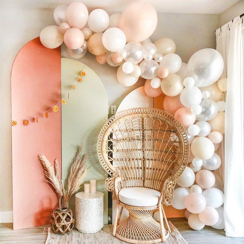 May include: A white wicker peacock chair with a white cushion sits in front of a wall decorated with a garland of white and peach balloons. The wall is painted in shades of peach and green.