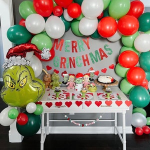 Pu&ograve; includere: Un palloncino verde Grinch con un cappello rosso, circondato da palloncini verdi, rossi e bianchi. Un tavolo con una tovaglia bianca &egrave; decorato con uno striscione che recita "Merry Grinchmas" e una ghirlanda di cuori rossi. Ci sono piccole figurine a tema Grinch e cupcake sul tavolo.
