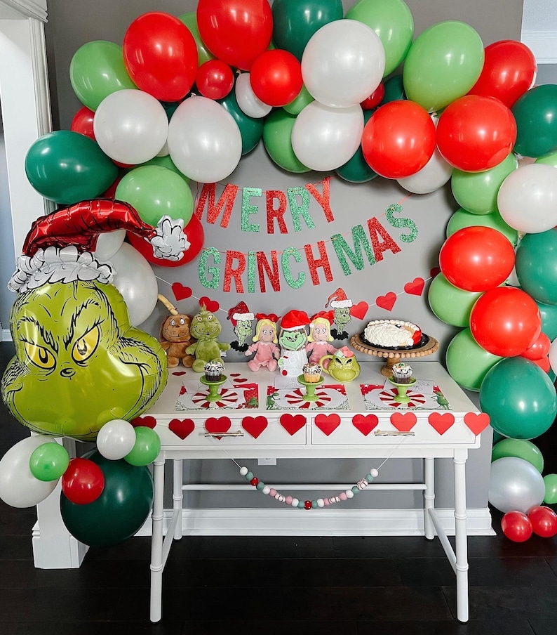 Pu&ograve; includere: Un palloncino verde Grinch con un cappello rosso, circondato da palloncini verdi, rossi e bianchi. Un tavolo con una tovaglia bianca &egrave; decorato con uno striscione che recita "Merry Grinchmas" e una ghirlanda di cuori rossi. Ci sono piccole figurine a tema Grinch e cupcake sul tavolo.
