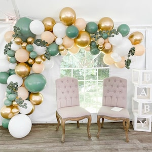 May include: A green, gold, and white balloon arch with greenery decorates a space with two pink chairs and a white block tower with the letters A, B, and Y.