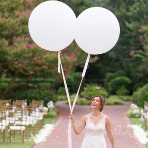 May include: A bride in a white wedding dress holds two large white balloons with pink ribbons. The balloons are floating in the air. The bride is smiling and looking up at the balloons.