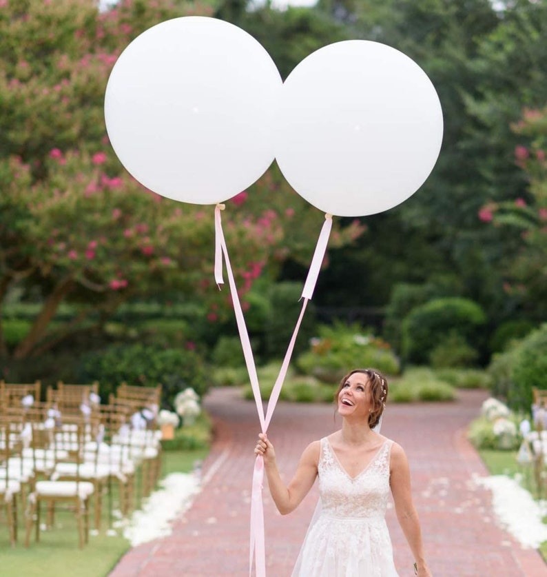 May include: A bride in a white wedding dress holds two large white balloons with pink ribbons. The balloons are floating in the air. The bride is smiling and looking up at the balloons.