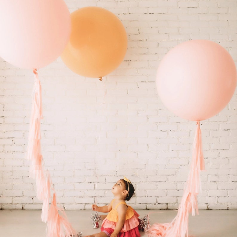 May include: A baby girl wearing a pink and orange dress sits in front of a white brick wall with three large balloons. The balloons are pink and peach with long pink tassels.