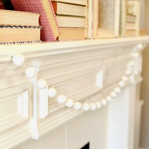 May include: A white pom pom garland draped across a white mantelpiece. The garland is made of small, fluffy white balls strung together on a white string.