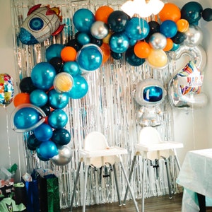 May include: A silver and blue balloon arch with orange and black balloons. The arch is decorated with a rocket and a silver astronaut balloon. The text "Happy Birthday" is on the astronaut balloon. Two white high chairs are in front of the arch.