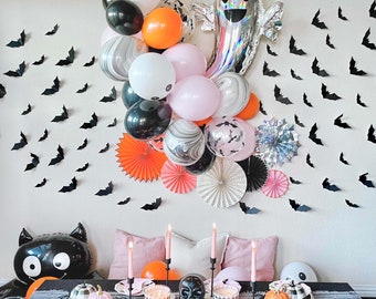 May include: A Halloween party table setting with a black and white ghost balloon, black and white bat cutouts, and a black cat balloon. The table is decorated with black, white, and orange balloons, and there are also some pumpkins and other Halloween decorations.