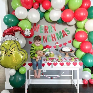Pu&ograve; includere: Un palloncino verde del Grinch con un cappello rosso, un bambino che tiene un peluche e un tavolo bianco con una ghirlanda di palloncini verdi e rossi, cupcake e una torta. Lo striscione recita "Merry Grinchmas".