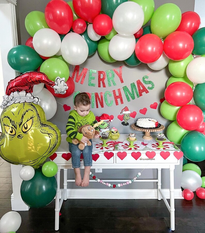 Pu&ograve; includere: Un palloncino verde del Grinch con un cappello rosso, un bambino che tiene un peluche e un tavolo bianco con una ghirlanda di palloncini verdi e rossi, cupcake e una torta. Lo striscione recita "Merry Grinchmas".