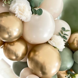 Puede incluir: Una colecci&oacute;n de globos dorados y blancos con follaje verde y flores blancas. Los globos est&aacute;n dispuestos en un racimo, creando un tel&oacute;n de fondo festivo y elegante.