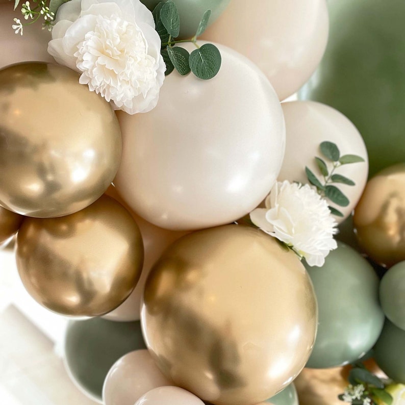 Puede incluir: Una colecci&oacute;n de globos dorados y blancos con follaje verde y flores blancas. Los globos est&aacute;n dispuestos en un racimo, creando un tel&oacute;n de fondo festivo y elegante.