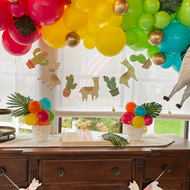 May include: A table setting for a party with a llama theme. The table is decorated with colorful balloons, llama cutouts, and cactus decorations. There are two vases with colorful balloons on the table.