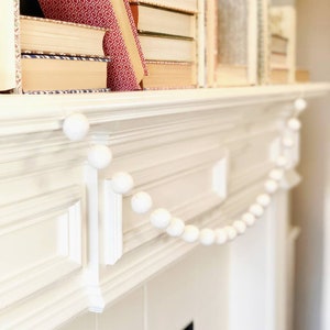 May include: A white fireplace mantel with a white felt ball garland draped across the top. The garland is made of small, round balls of felt, strung together on a white string.