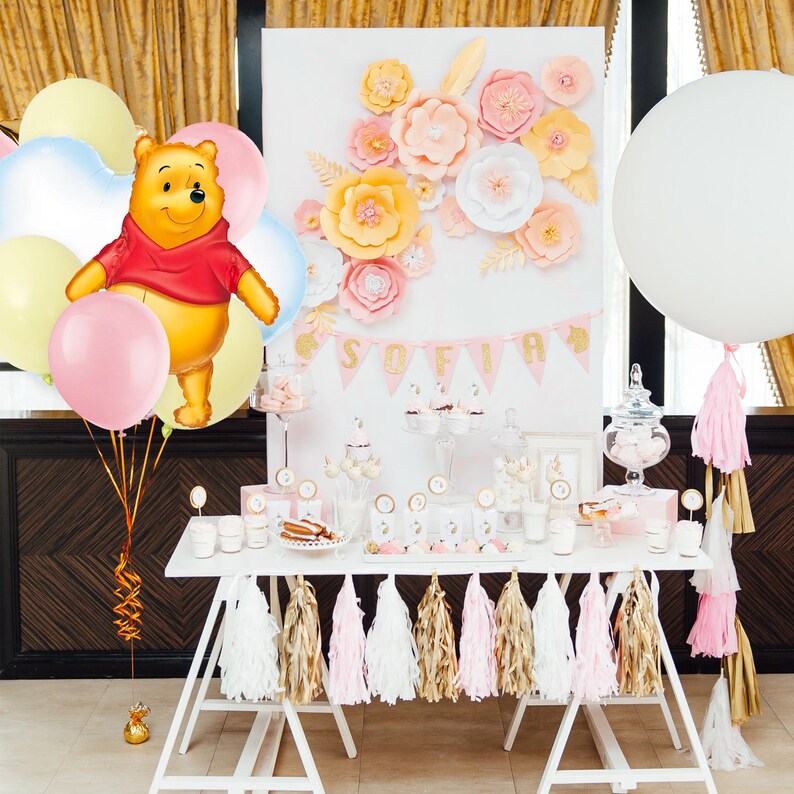 May include: A white table with a pink and gold dessert table decorated with paper flowers, balloons, and a Winnie the Pooh character. The table is decorated with pink, white, and gold tassel garlands. The table is set up for a party with a sign that says "Sofia".
