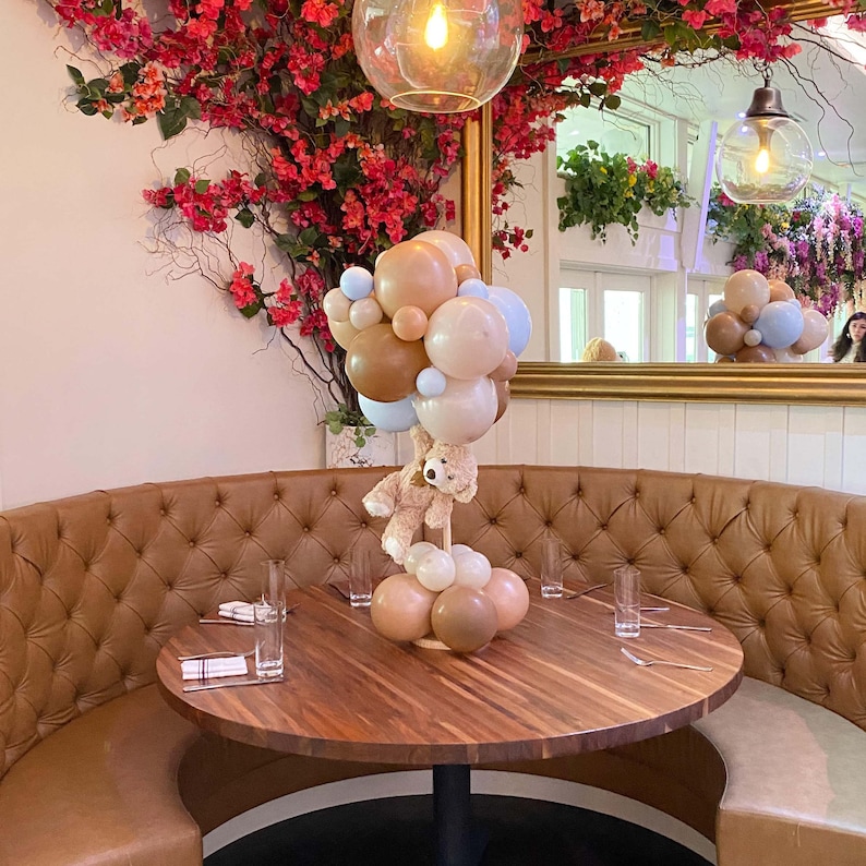 Op de afbeelding: Een bruine leren booth met een ronde houten tafel in het midden. De tafel is versierd met een middelpunt van ballonnen in tinten bruin en blauw, met een teddybeer die de ballonnen vasthoudt. Twee glazen en een servet staan op de tafel.