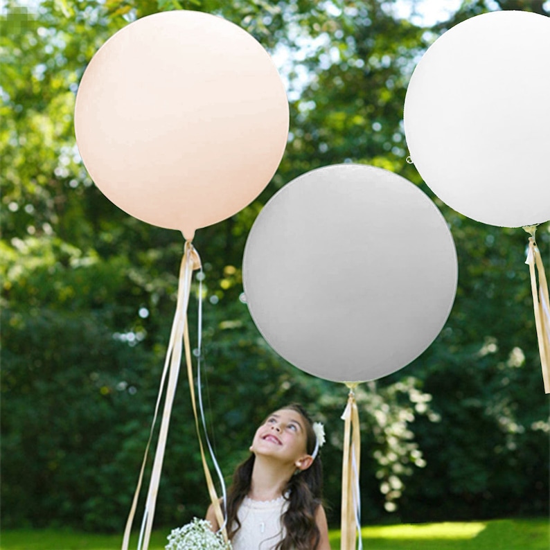 May include: Three large, round balloons in shades of white, light gray, and blush pink. The balloons are tied with long, thin ribbons and are floating in the air. A young girl is standing in front of the balloons, looking up at them.