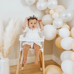 May include: A baby girl wearing a white dress and a flower crown sits in a white high chair with wooden legs. The background is decorated with a garland of white and cream balloons.