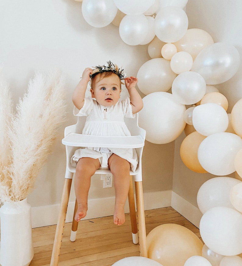 May include: A baby girl wearing a white dress and a flower crown sits in a white high chair with wooden legs. The background is decorated with a garland of white and cream balloons.