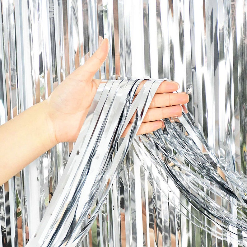 May include: Silver tinsel curtain with a hand holding a few strands of the curtain. The curtain is made of thin strips of silver foil.